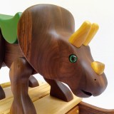 hand carved wooden triceratops dinosaur rocking toy. The dino is carved from walnut wood, left natural with oil finish. Poplar hand carved horns. It has a green painted wood saddle and leather handle. Green reptile eyes. Walnut rockers with maple slats. Small, size for 1-2 years old.