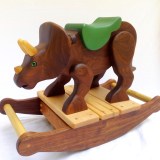 hand carved wooden triceratops dinosaur rocking toy. The dino is carved from walnut wood, left natural with oil finish. Poplar hand carved horns. It has a green painted wood saddle and leather handle. Green reptile eyes. Walnut rockers with maple slats. Small, size for 1-2 years old.