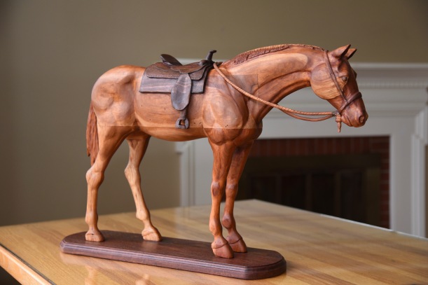 hand carved cherry wood horse standing with carved walnut saddle on walnut base. With carved bridle and rein over neck
