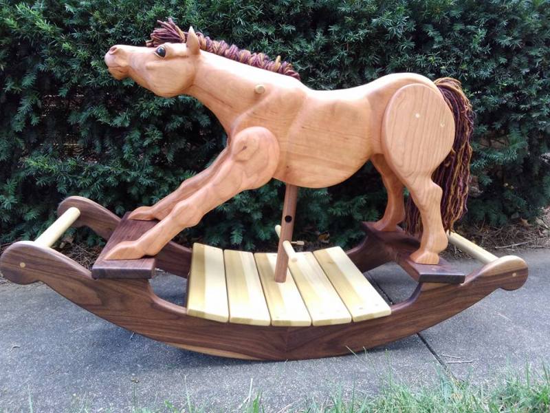 hand carved rocking horse in cherry wood that is stretching out its front legs and looking up. multicolored yarn mane and tail. all natural wood construction. background of green leaves.