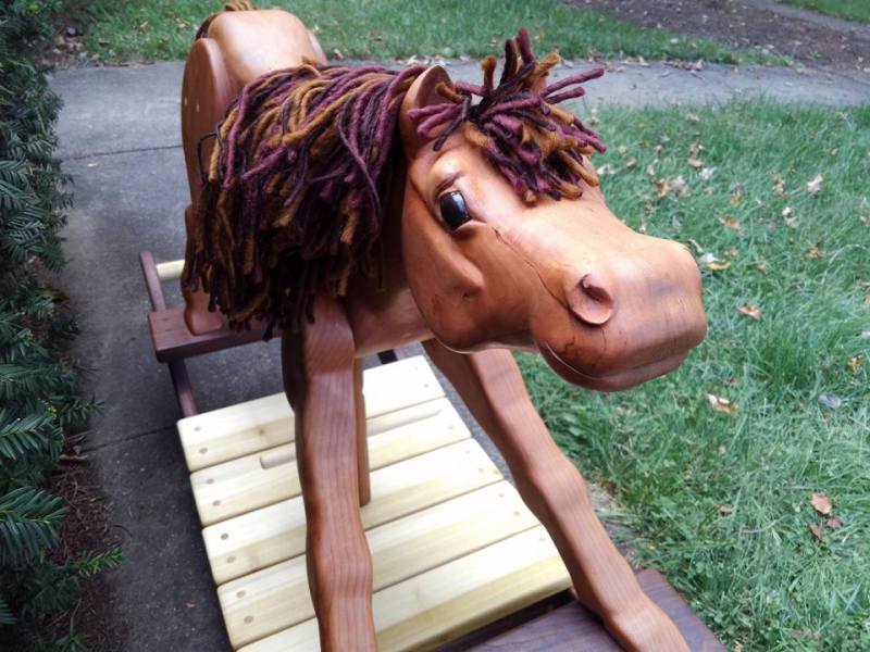 Hand carved wooden rocking horse in cherry wood with walnut and poplar rockers. hand painted eyes create a cute happy expression. horse pictured on sidewalk with background of grass and green leaves.